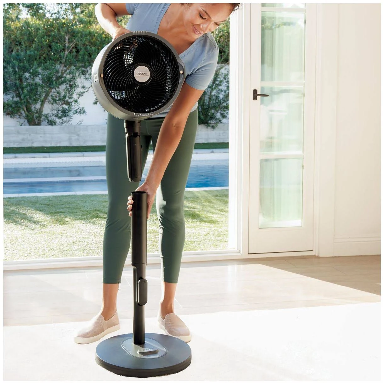 Mujer ensamblando un ventilador negro y gris. Usa ropa deportiva. El fondo tiene piscina, césped verde y una ventana.