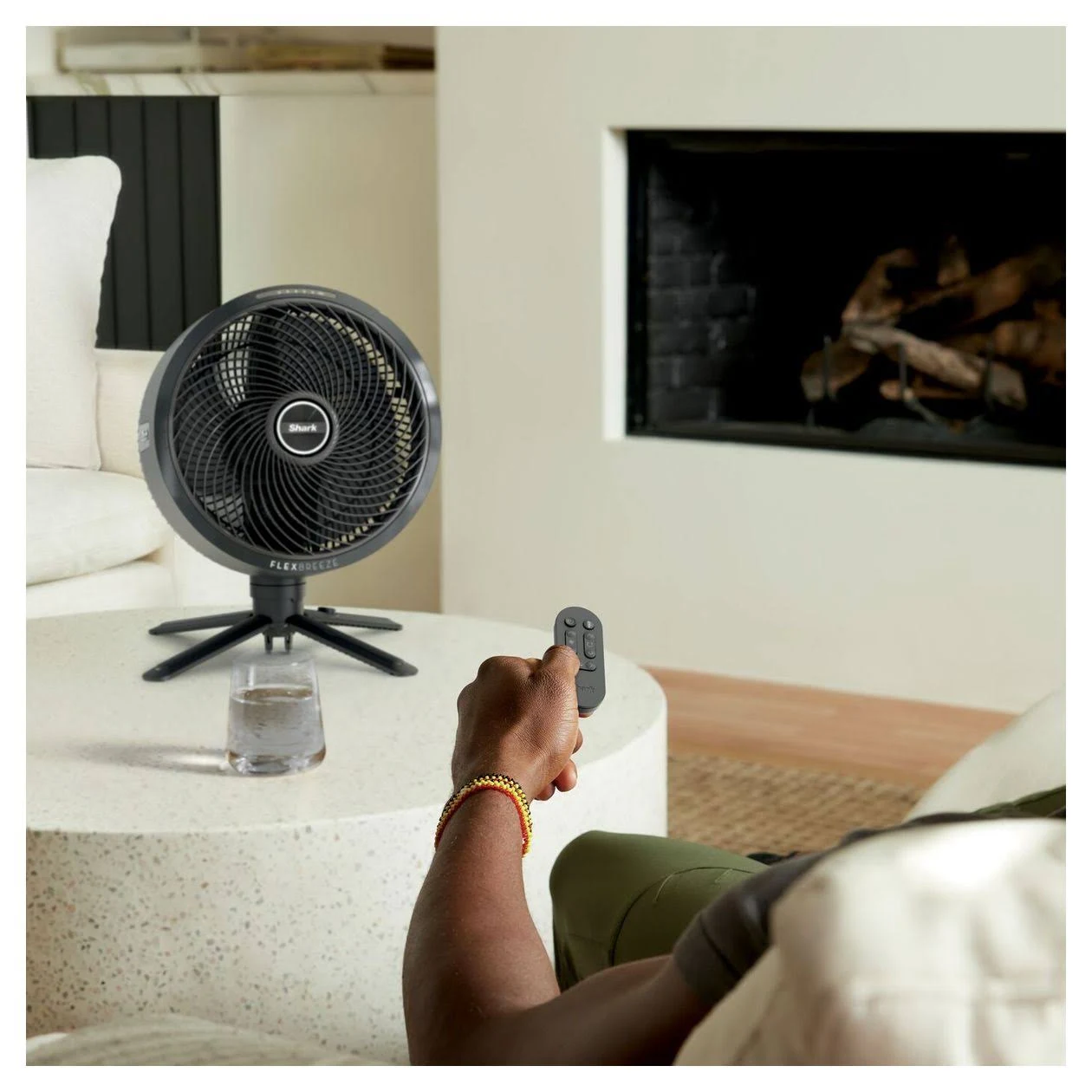 Una persona con un mando a distancia controla un ventilador negro sobre una mesa. Un vaso de agua está cerca. En el fondo se ve una chimenea.