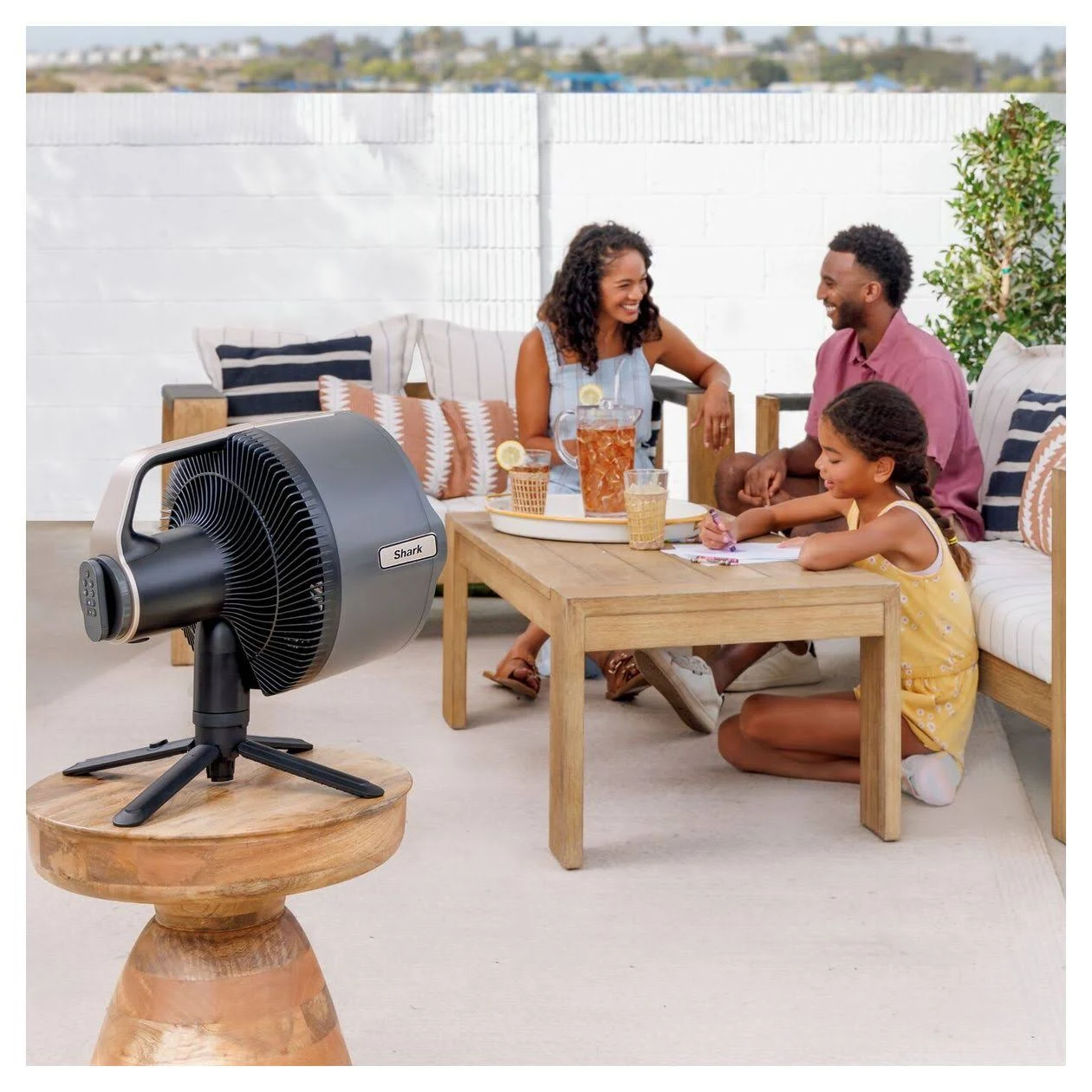 Una familia reunida al aire libre. Un ventilador sobre una mesa de madera. Una mujer, un hombre y una niña sentados, con bebidas y materiales de dibujo.
