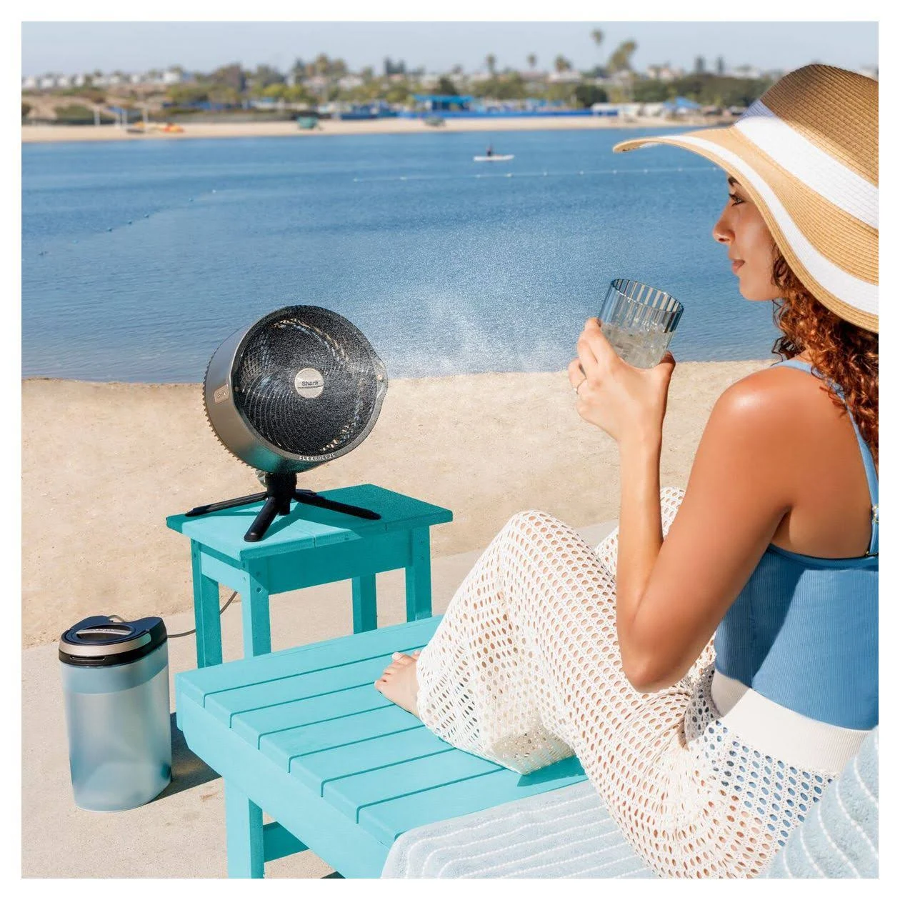 Mujer en la playa con un ventilador y un vaso de agua. Lleva sombrero y pantalones blancos. Agua azul y playa al fondo.