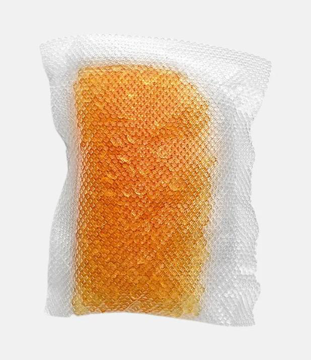 Ein orangefarbenes Gelpack ist in einem weißen, strukturierten, netzartigen Material eingeschlossen. Der Hintergrund ist ein schlichtes, hellgraues.