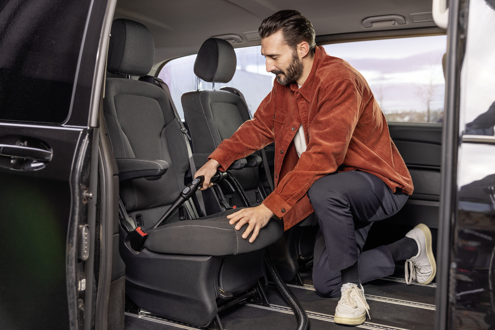 Mężczyzna odkurza siedzenie samochodu. Ma na sobie czerwoną koszulę i ciemne spodnie. Wnętrze samochodu jest czarne.