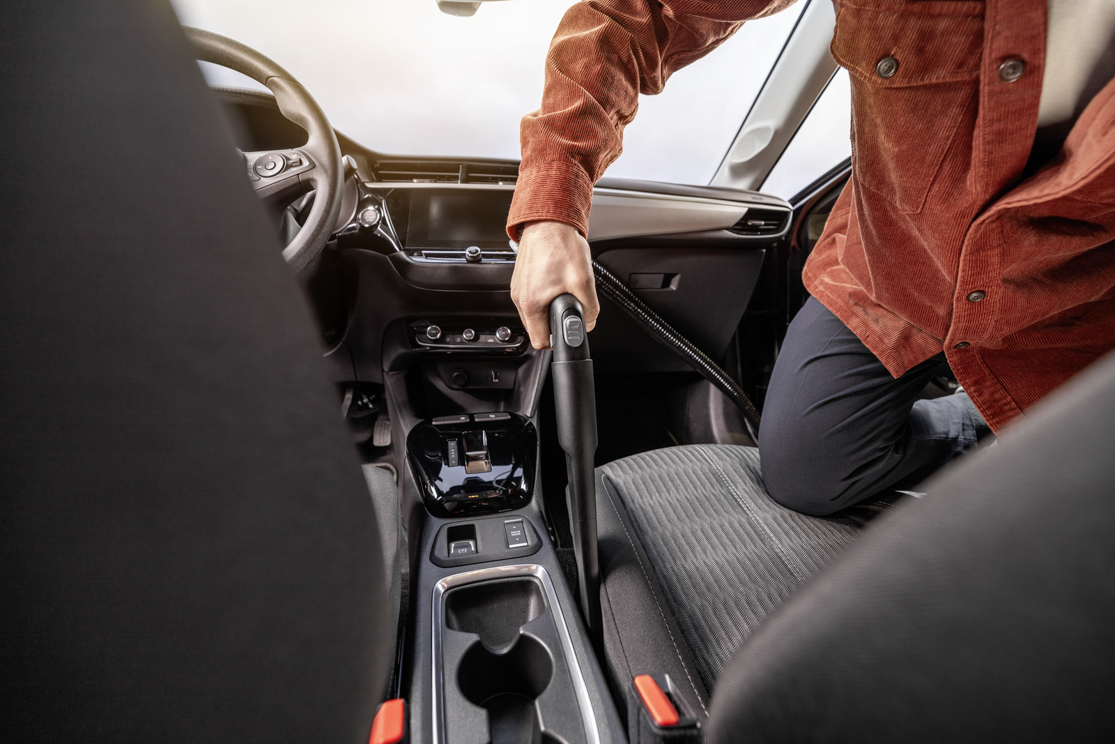 Osoba odkurza wnętrze samochodu. Pomarańczowa kurtka, czarne spodnie. Widoczna kierownica, deska rozdzielcza i siedzenia. Odkurzacz w użyciu.