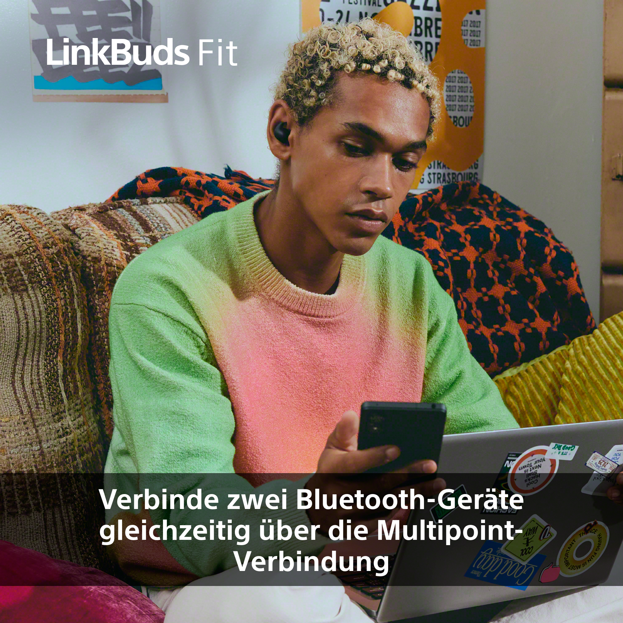 Eine Person mit Kopfhörern hält ein Telefon und sitzt auf einem Sofa. Im Hintergrund sind Laptop und Poster zu sehen.