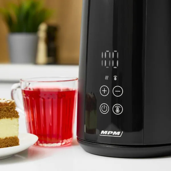 Czarny czajnik elektryczny z cyfrowym wyświetlaczem pokazuje 100. Kubek czerwonego płynu, kawałek ciasta i roślina w tle.