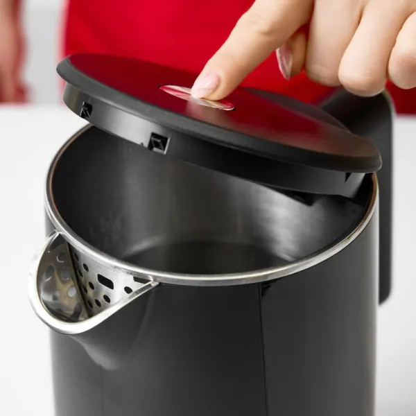 Ręka naciska przycisk na pokrywie czarnego czajnika. Czajnik jest czarny i srebrny. Czerwona koszula w tle.