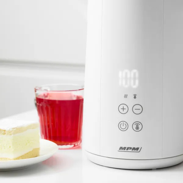 Biały czajnik elektryczny pokazuje 100 stopni. Szklanka czerwonego soku i kawałek ciasta leżą na białym stole.