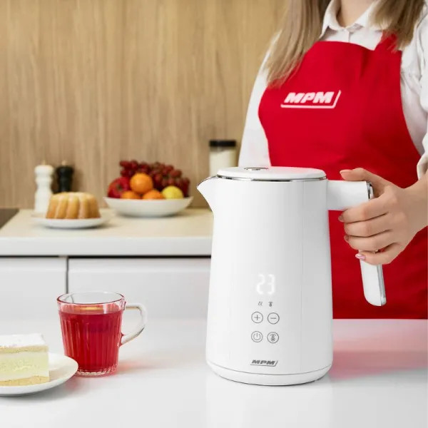 Biały czajnik elektryczny z cyfrowym wyświetlaczem na blacie kuchennym. Kobieta w czerwonym fartuchu go trzyma. Przed nim szklanka herbaty.