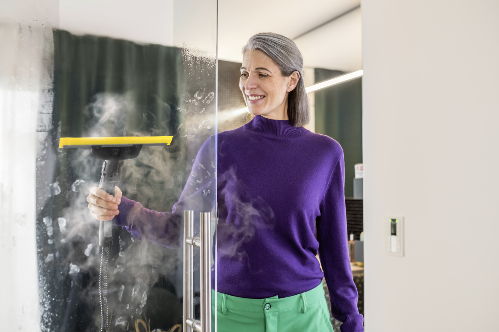 Kobieta czyści szklane drzwi za pomocą parownicy. Ma na sobie fioletowy sweter i zielone spodnie. Para wypełnia powietrze.