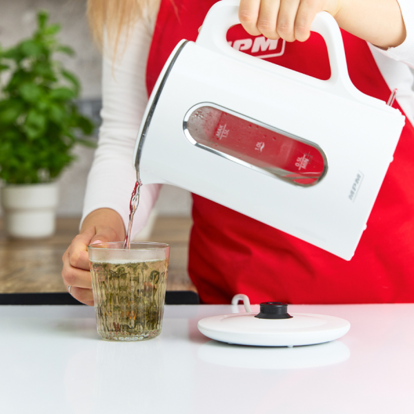 Kobieta wlewa gorącą wodę z białego czajnika do szklanki z ziołami. Widoczny jest czerwony fartuch, biały stół i roślina.