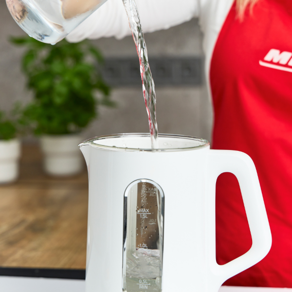 Woda wlewa się ze szklanki do białego czajnika elektrycznego. Widoczny jest czerwony fartuch. Rośliny i drewniana deska w tle.