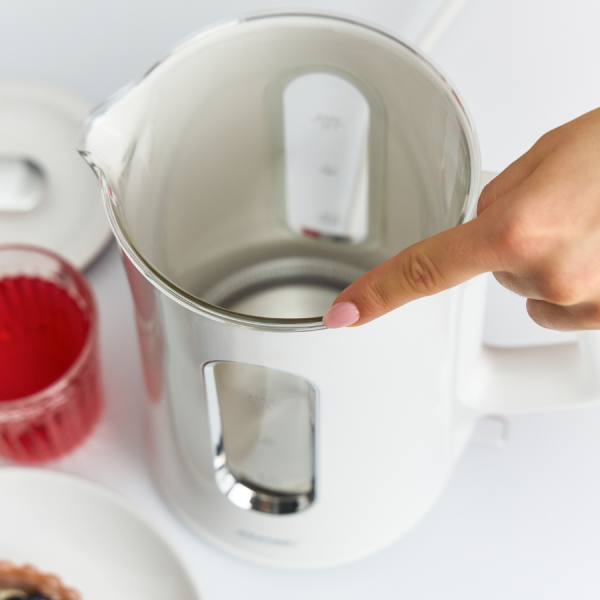 Ręka wskazuje na krawędź białego czajnika elektrycznego. Szklanka czerwonego płynu i tarty znajdują się w tle na białej powierzchni.