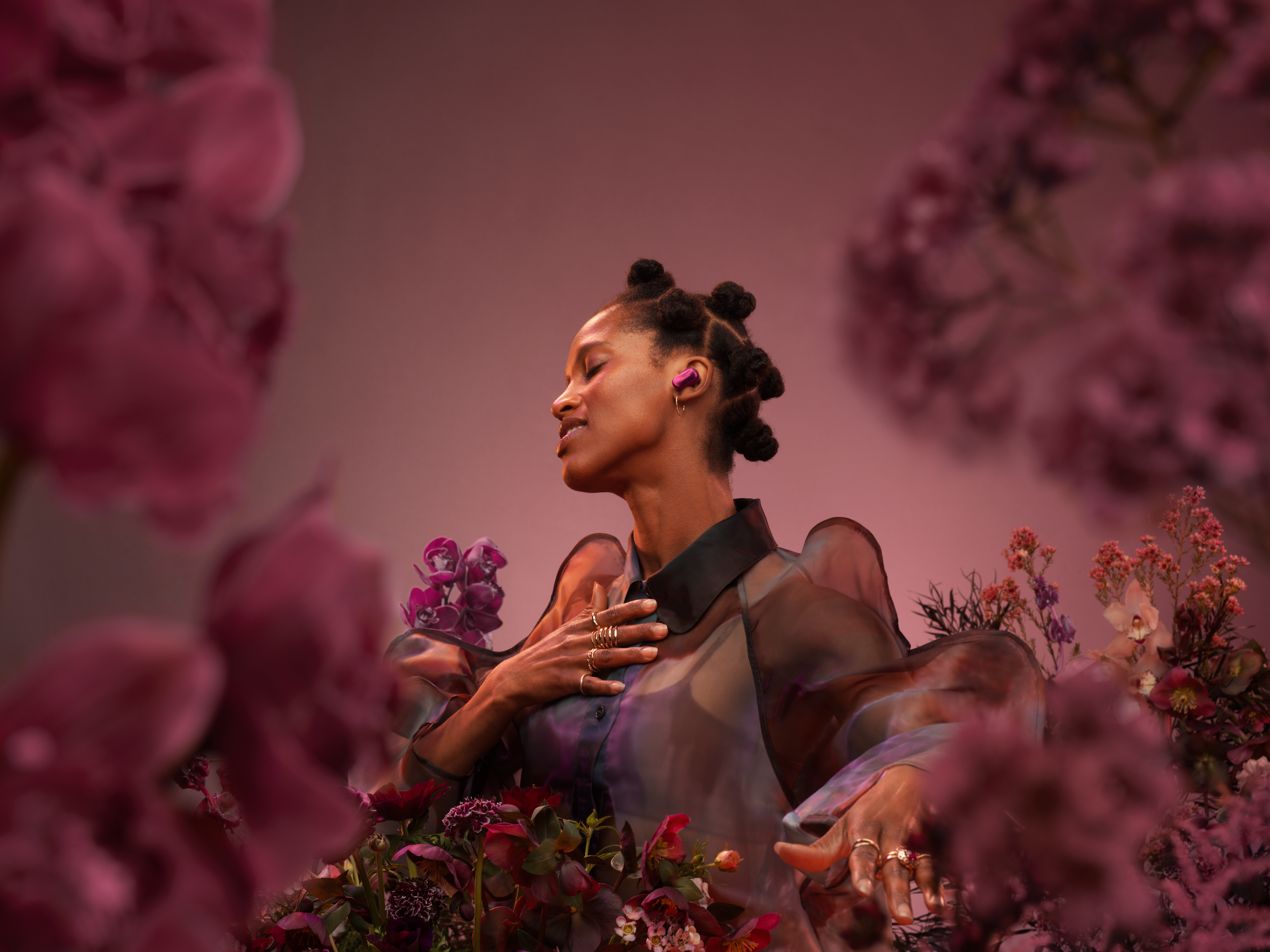 Une femme portant des écouteurs et des bagues roses, entourée de fleurs roses. Elle porte une chemise noire transparente, les yeux fermés.