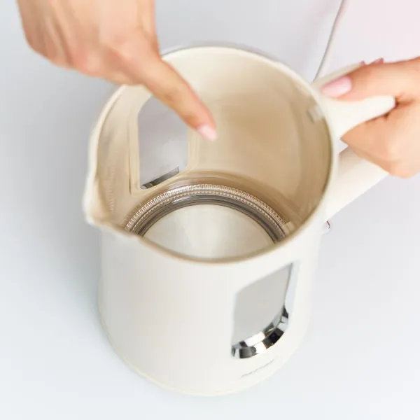 Kremowy czajnik elektryczny jest otwarty, palec wskazuje do środka. Czajnik stoi na białej powierzchni.