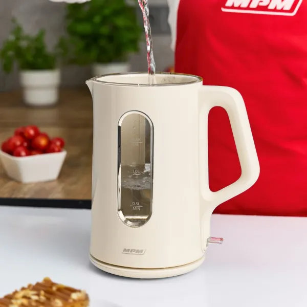 Kremowy czajnik elektryczny jest napełniany wodą. Osoba w czerwonym fartuchu stoi obok. Pomidory i rośliny w tle.