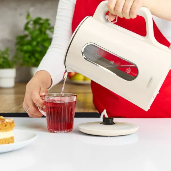 Osoba wlewa gorącą wodę z kremowego czajnika do szklanki z czerwonym płynem. Blisko plasterek ciasta i pokrywka czajnika.
