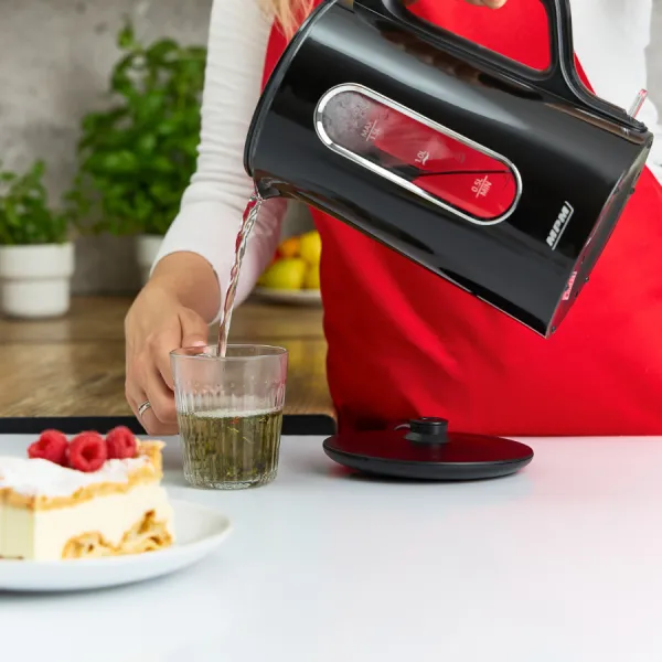 Osoba wlewa wodę z czarnego czajnika do szklanki herbaty. Kawałek ciasta z malinami leży na talerzu.