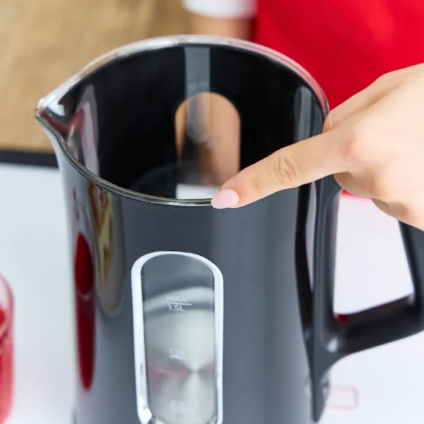 Czarny czajnik elektryczny z dłonią wskazującą jego krawędź. Czajnik ma wskaźnik poziomu wody. Tło to rozmyte czerwone ubrania.