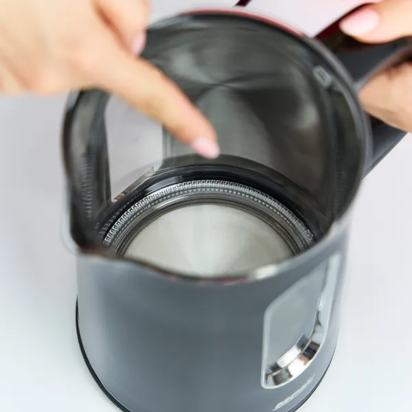 Ręka wskazuje do wnętrza czarnego czajnika elektrycznego. Wnętrze czajnika jest widoczne. Tło jest białe.