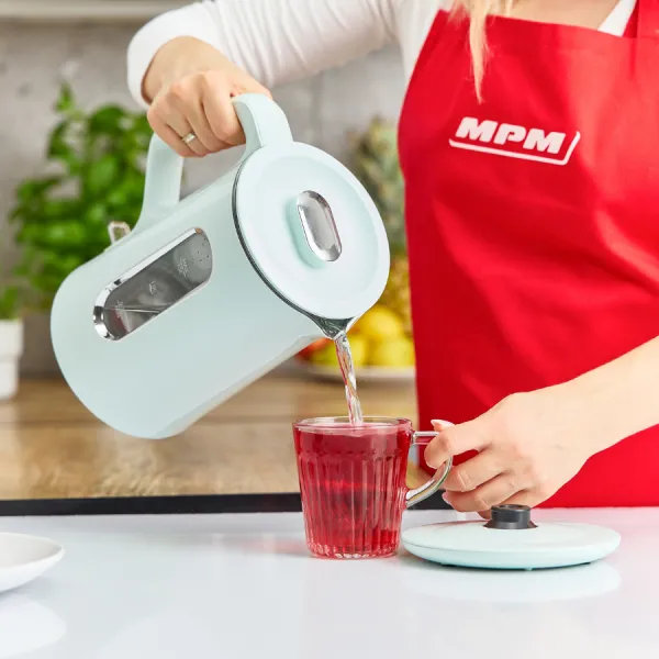 Osoba wlewa gorącą wodę z jasnoniebieskiego czajnika do szklanki czerwonej herbaty, w czerwonym fartuchu.