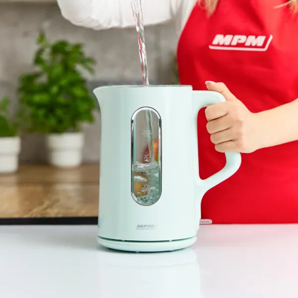 Woda wlewana do jasno-niebieskiego czajnika. Osoba w czerwonym fartuchu trzyma czajnik. Rośliny w tle.