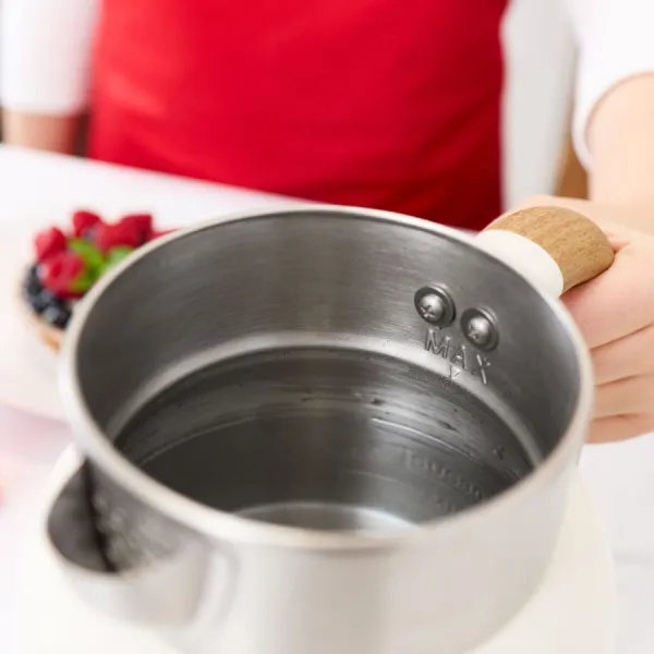 Osoba trzyma czajnik ze stali nierdzewnej. Deser z jagodami w tle. Widoczny czerwony fartuch.