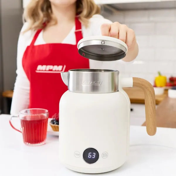 Kobieta otwiera biały czajnik elektryczny z drewnianym uchwytem. Czerwony fartuch, filiżanka herbaty i deser są w scenie.