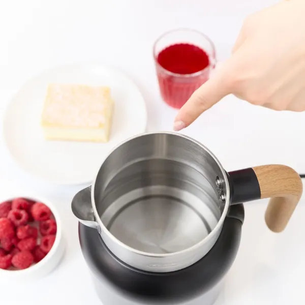 Ręka wskazuje na czajnik ze stali nierdzewnej z drewnianym uchwytem. W pobliżu maliny, ciasto i czerwony napój na białej powierzchni.