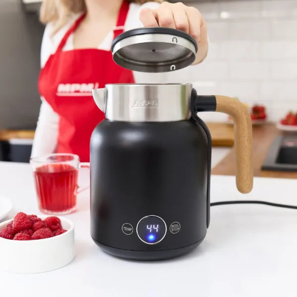 Kobieta podnosi pokrywkę czarnego czajnika z drewnianą rączką. Miska malin i szklanka czerwonego płynu na stole.