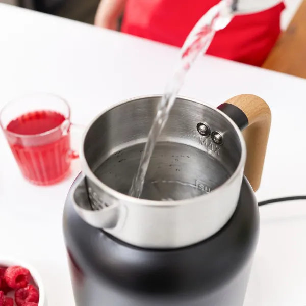 Woda wlewana do czajnika. Szklanka czerwonego płynu i maliny są w pobliżu. Osoba w czerwonym fartuchu jest w tle.