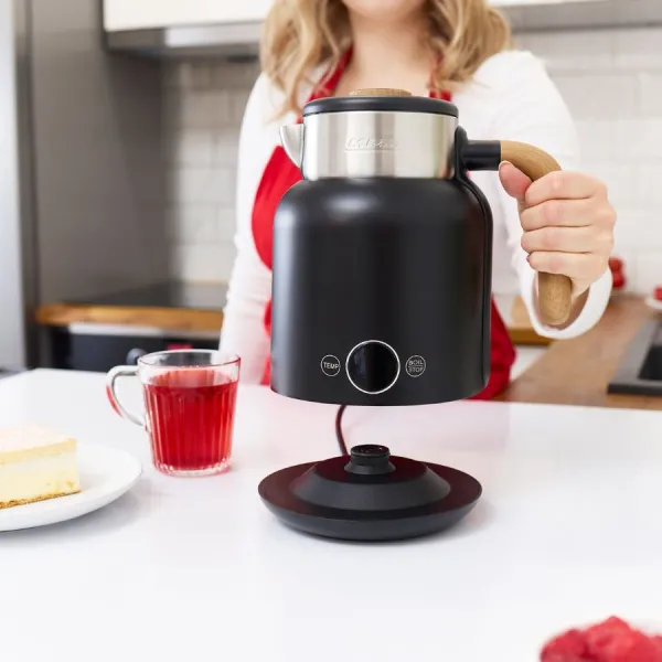 Kobieta trzyma czarny czajnik elektryczny z drewnianym uchwytem. Na stole szklanka czerwonego płynu, kawałek ciasta i maliny.