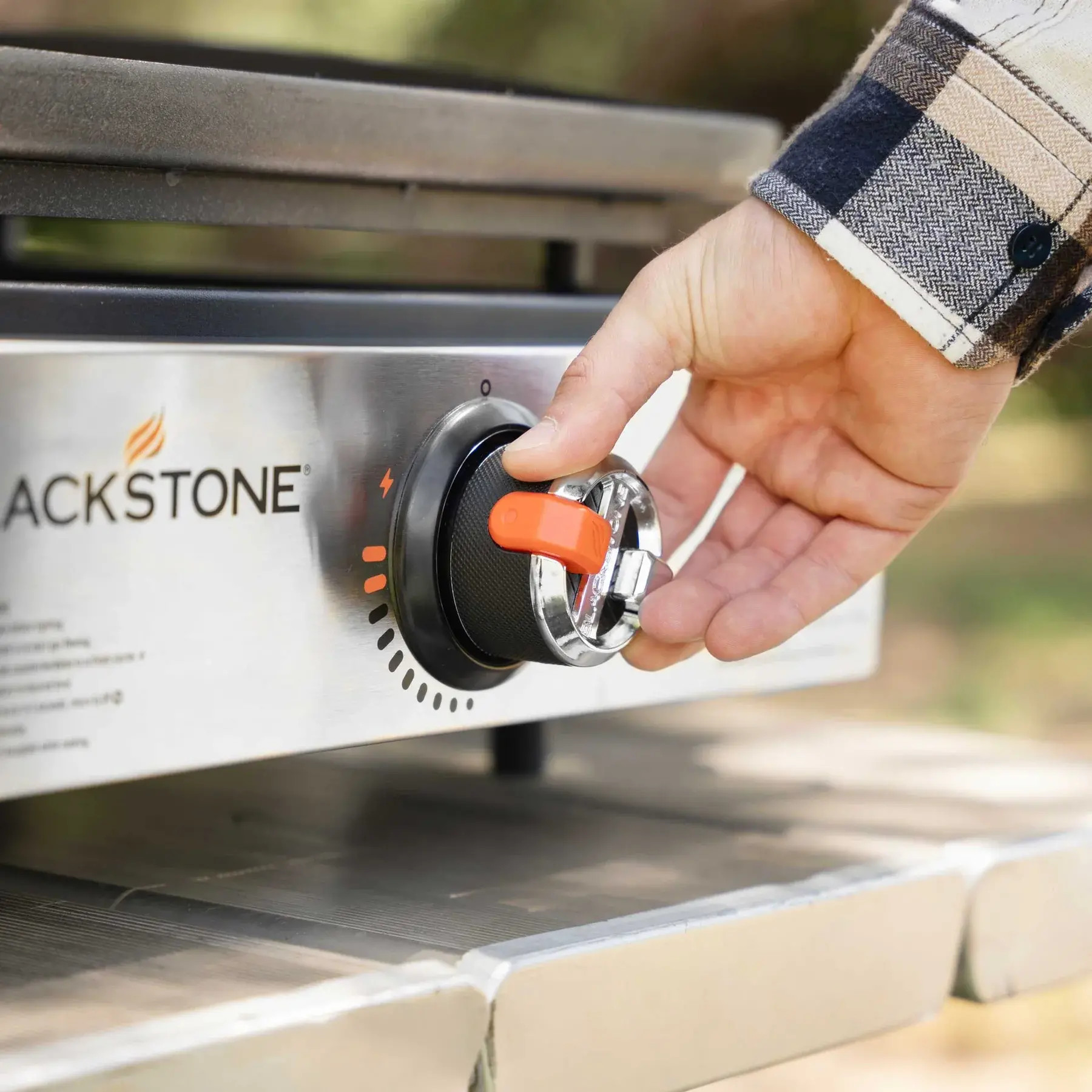 Ręka przekręca pokrętło grilla. Czarne pokrętło ma pomarańczowy wskaźnik. Grill jest srebrny z logo Blackstone.