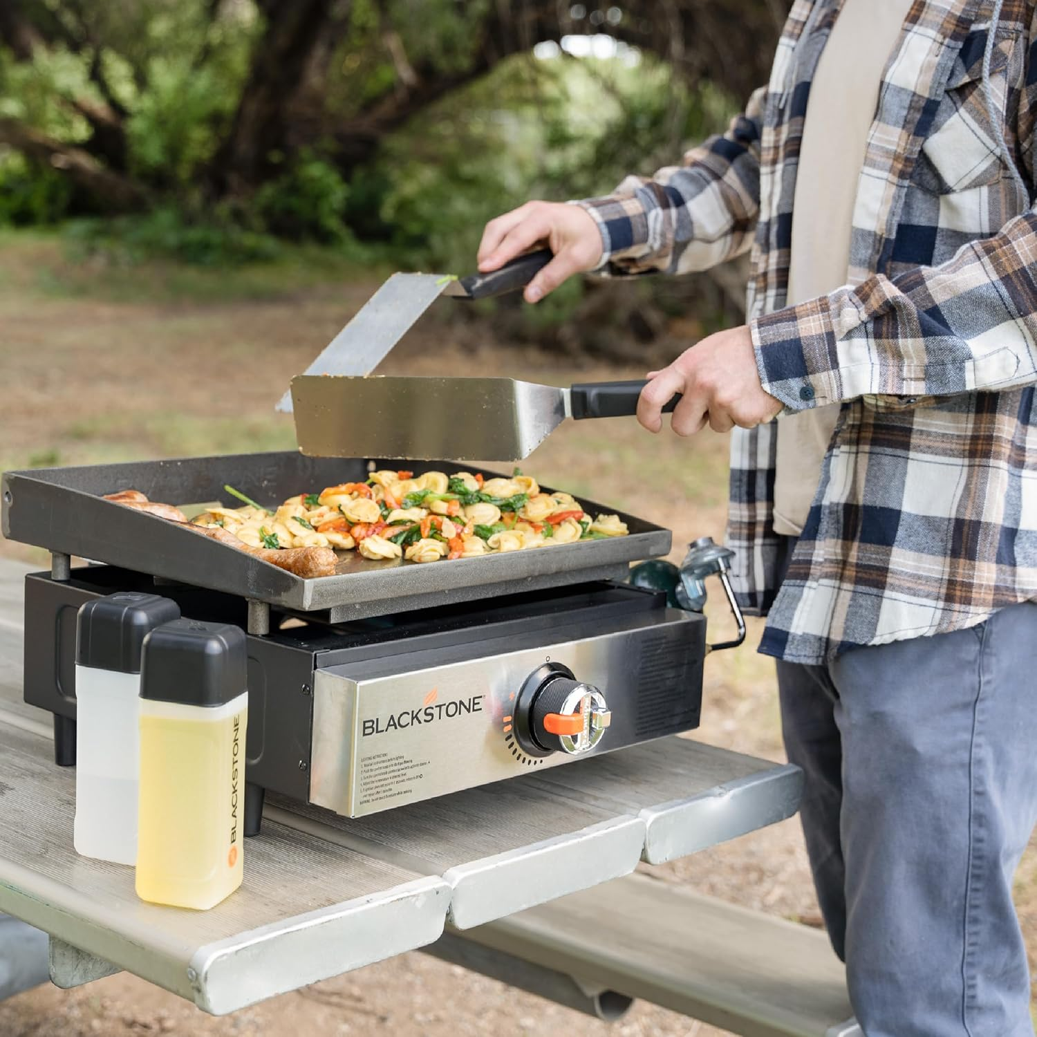 Osoba gotuje jedzenie na grillu Blackstone na stole piknikowym. Ma na sobie koszulę w kratę. Na stole są dwie butelki.