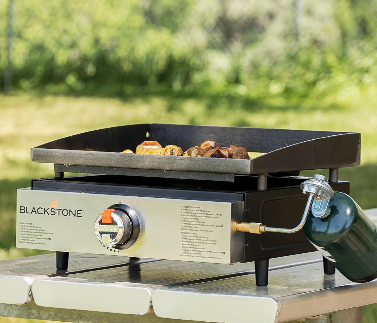 Grill Blackstone z jedzeniem, zbiornikiem propanu i metalowym stołem. Tło to rozmyta zielona trawa i drzewa.