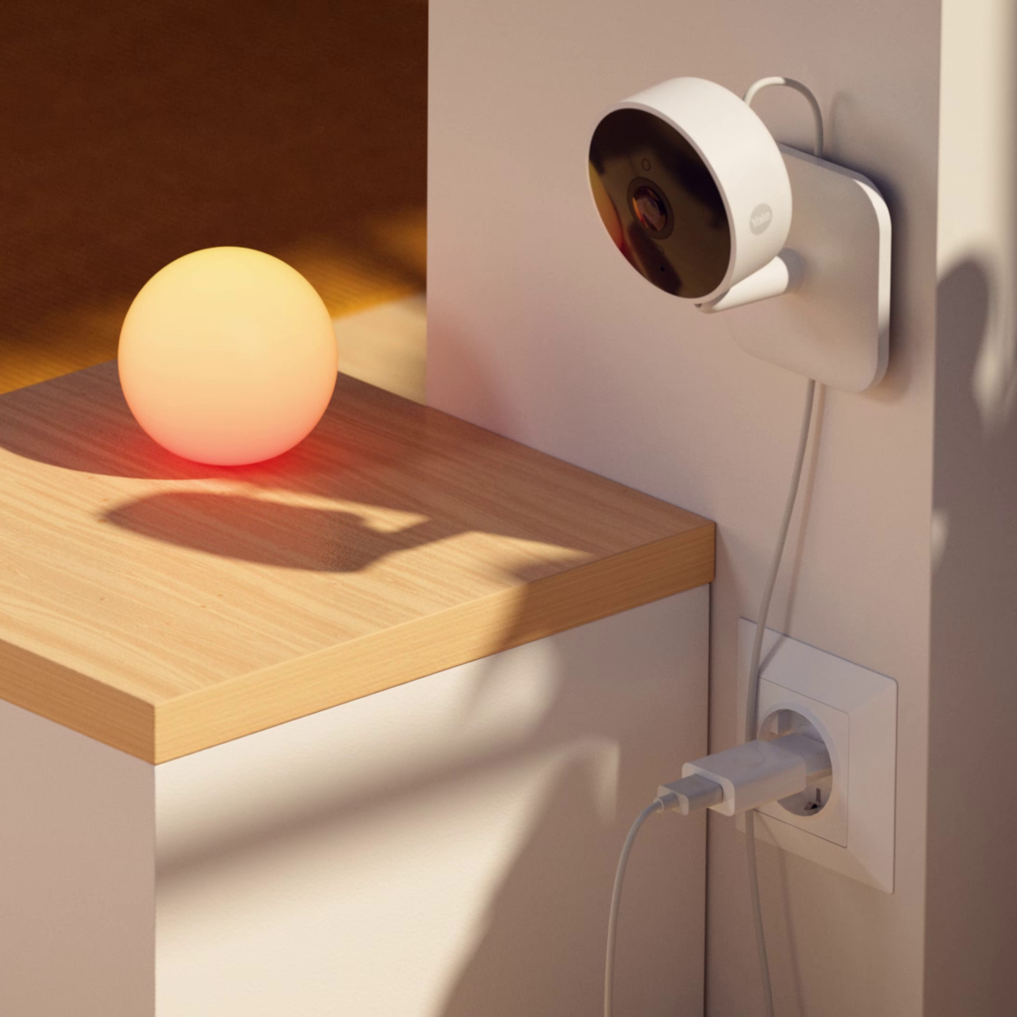 Eine weiße Überwachungskamera ist an der Wand montiert. Eine leuchtende Kugel-Lampe steht auf einem Holztisch. Ein Stecker steckt in der Wand.