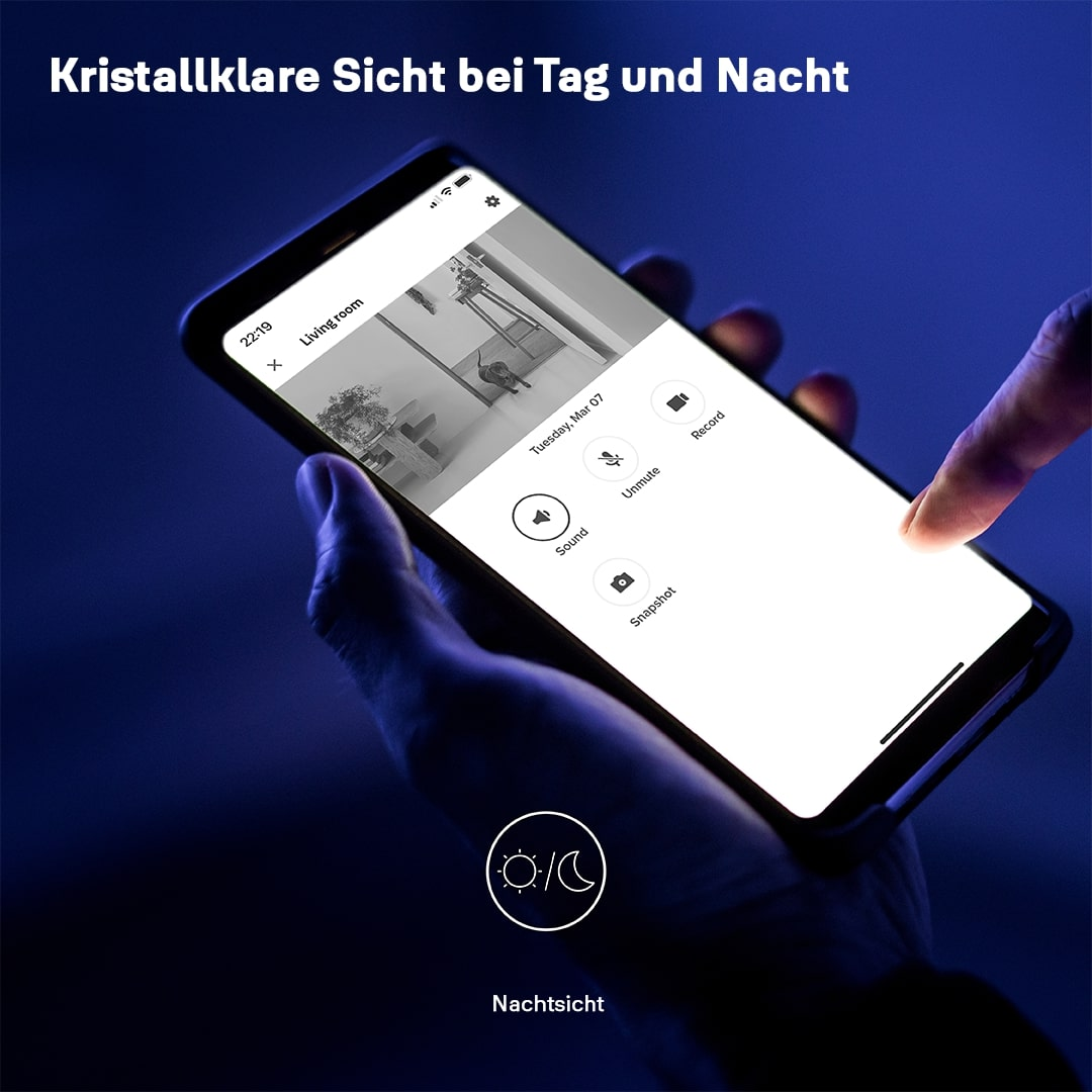 Eine Hand hält ein Telefon, das einen Live-Video-Feed des Wohnzimmers zeigt. Der Bildschirm zeigt ein Zimmer mit Möbeln. Ein Finger berührt den Bildschirm.