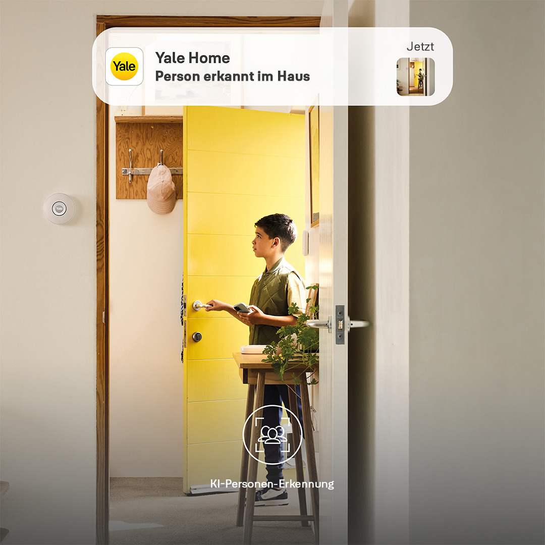 Ein Junge steht in einer Tür, schaut auf ein Telefon. Hinter ihm eine gelbe Tür. Oben ein Yale Home Logo.