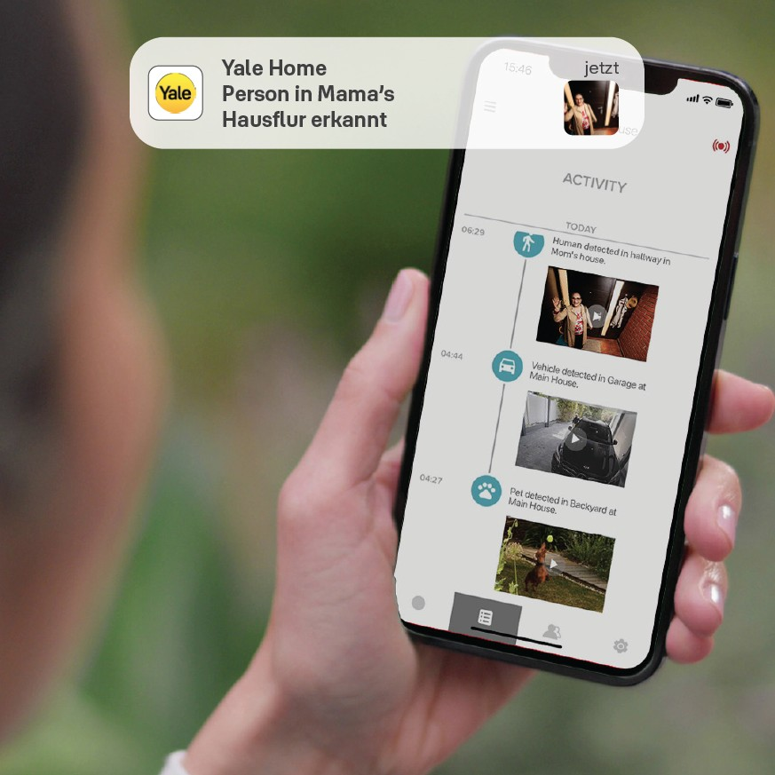 Eine Person hält ein Telefon mit geöffneter Yale Home App. Der Bildschirm zeigt Aktivitätsprotokolle. Ein Alarm sagt: "Person in Mama's Hausflur erkannt".