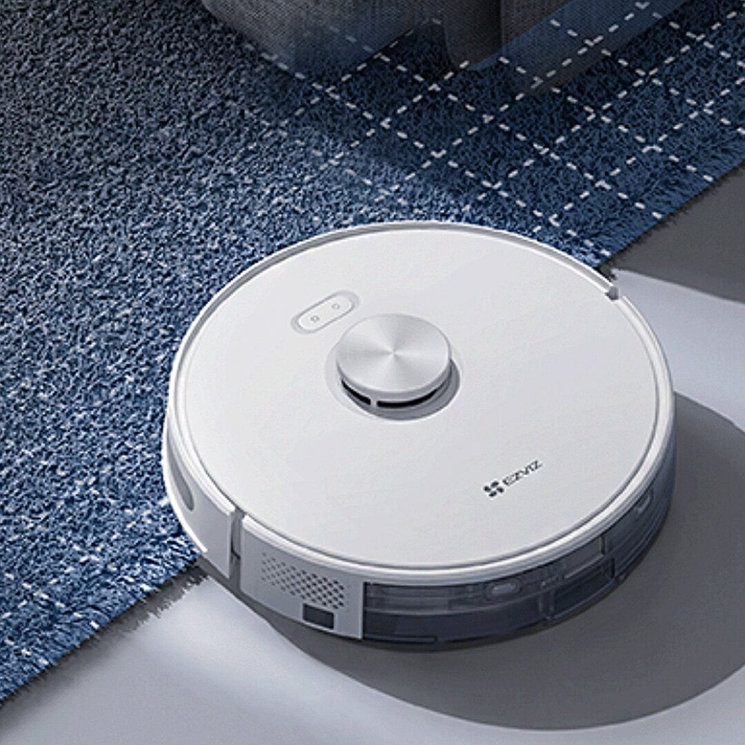 Biały odkurzacz automatyczny na niebieskim dywanie. Ma srebrny wierzch i znajduje się obok wzorzystego dywanu i szarej powierzchni.