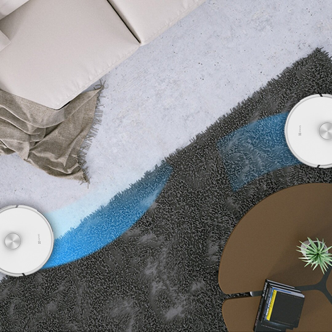 Dwa białe roboty odkurzacze czyszczą ciemny dywan. Widać również sofę i stolik kawowy.