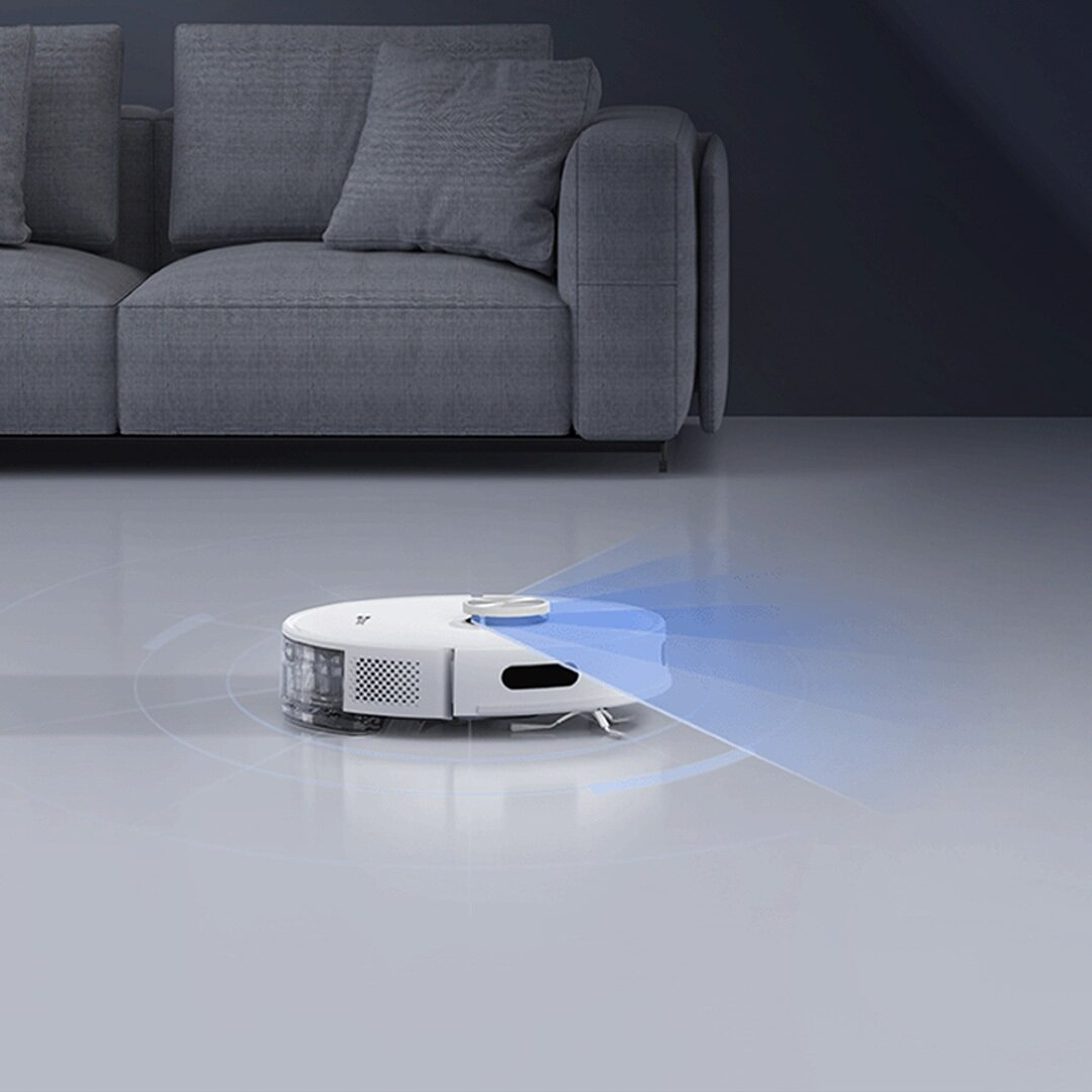 Biały odkurzacz robot na jasnoszarej podłodze. Szara sofa w tle. Odkurzacz emituje niebieskie światło.