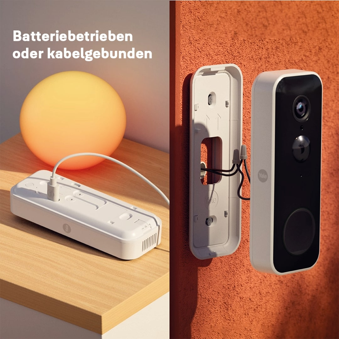 Eine weiße Türklingel ist an einer orangefarbenen Wand montiert. Eine Ladestation und eine orangefarbene Kugel befinden sich auf der linken Seite. Der Text lautet 'Batterie oder Kabel'.