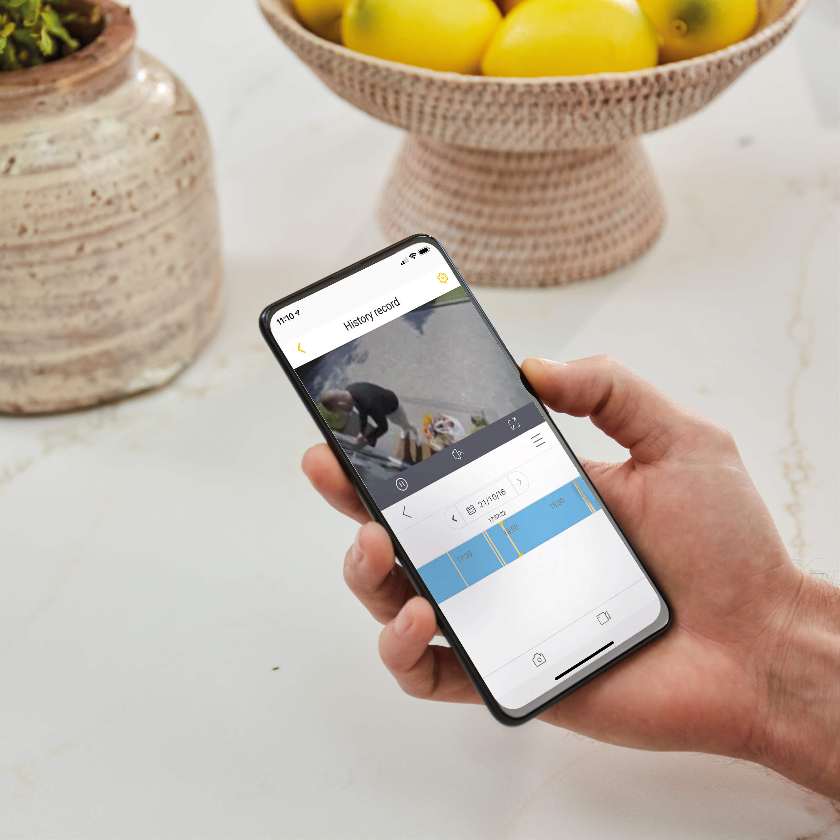 Eine Hand hält ein Telefon, das einen Verlaufsdatensatz anzeigt. Zitronen in einem Korb sind im Hintergrund. Links ist eine Vase.