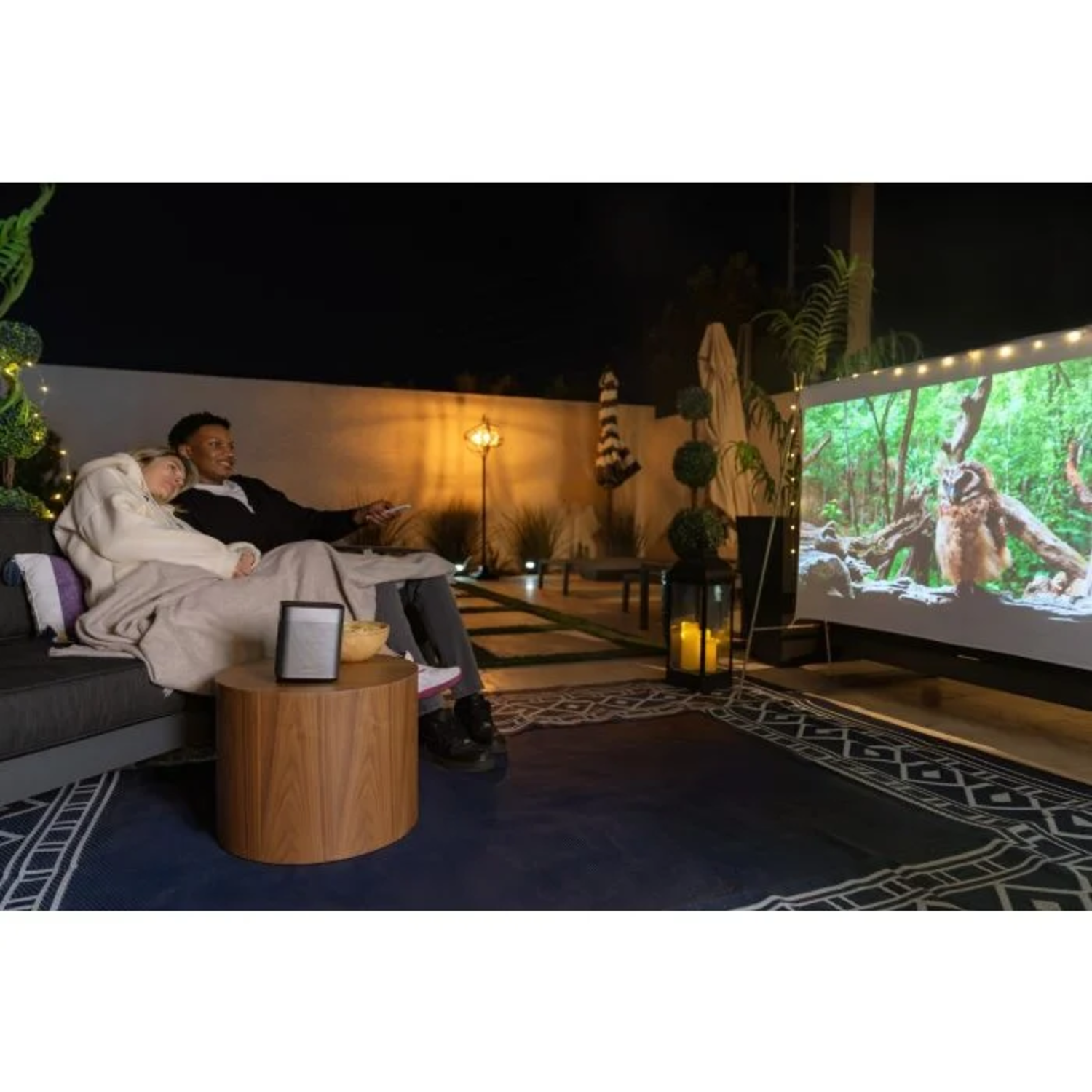 Couple watching a movie outdoors at night. Projector screen with nature scene. Wooden table with speaker, food and drinks.
