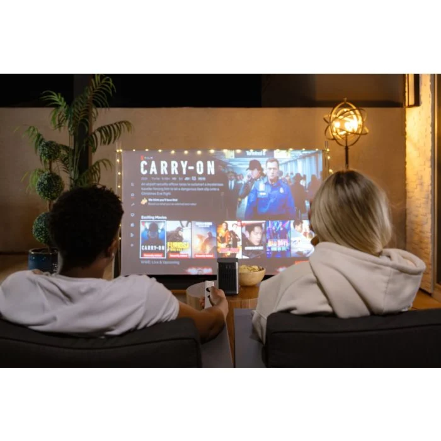 Two people watch a movie projected on a screen outside. A small projector sits near them, and a lamp is to the right.