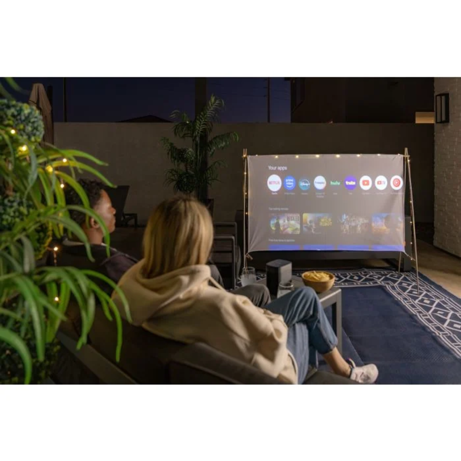 Two people watching a movie on a projector screen outdoors. Snacks and drinks on a table in front of them.