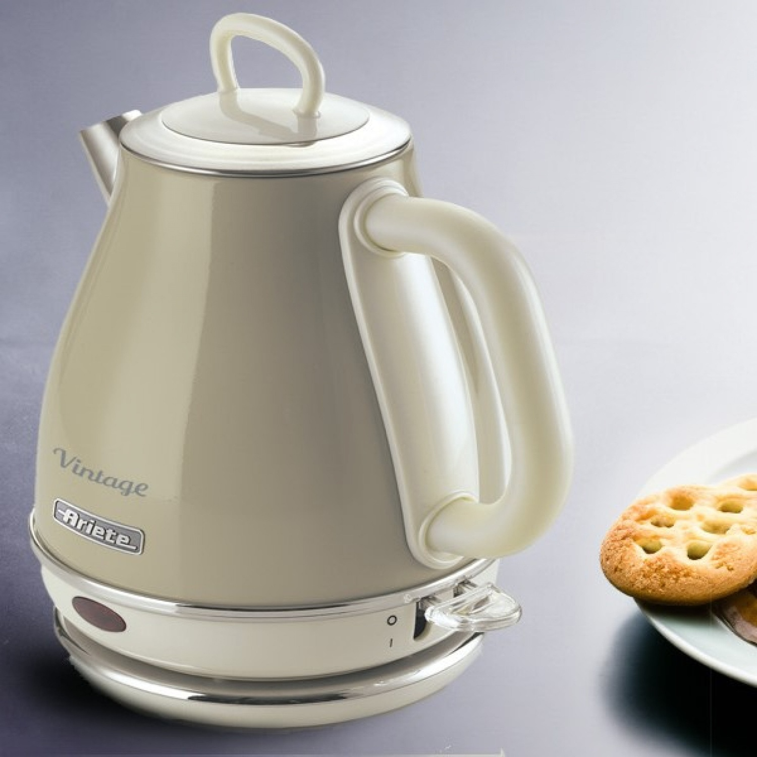 Bollitore elettrico color crema con manico e coperchio bianchi, accanto a un piatto di biscotti. Sfondo grigio chiaro.
