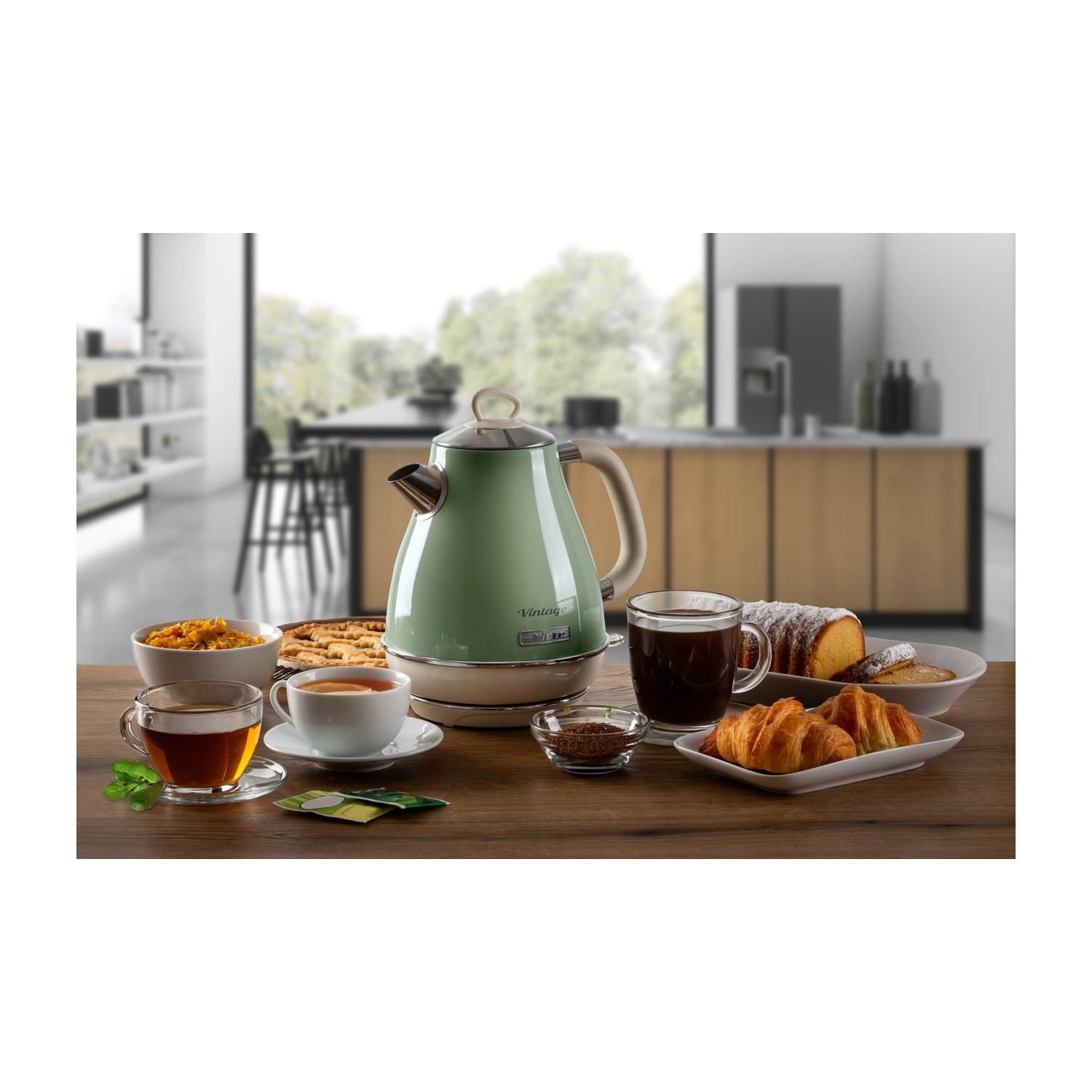 Un tavolo con tè, caffè, pasticcini e un bollitore verde. Lo sfondo mostra una cucina con una finestra.