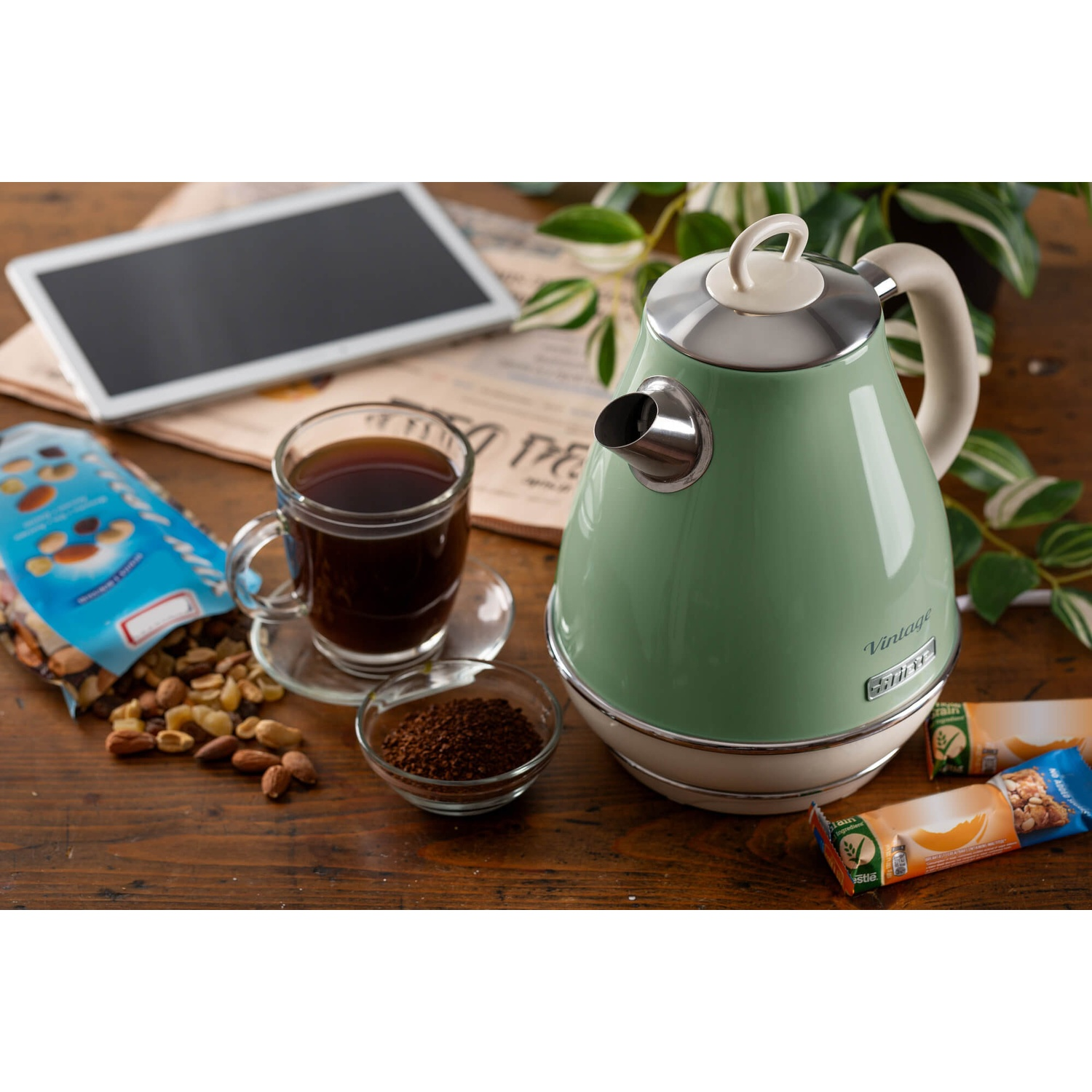 Un tavolo con un bollitore verde, caffè, noci e un tablet. Sono presenti anche una tazza di caffè e una ciotola di fondi di caffè.