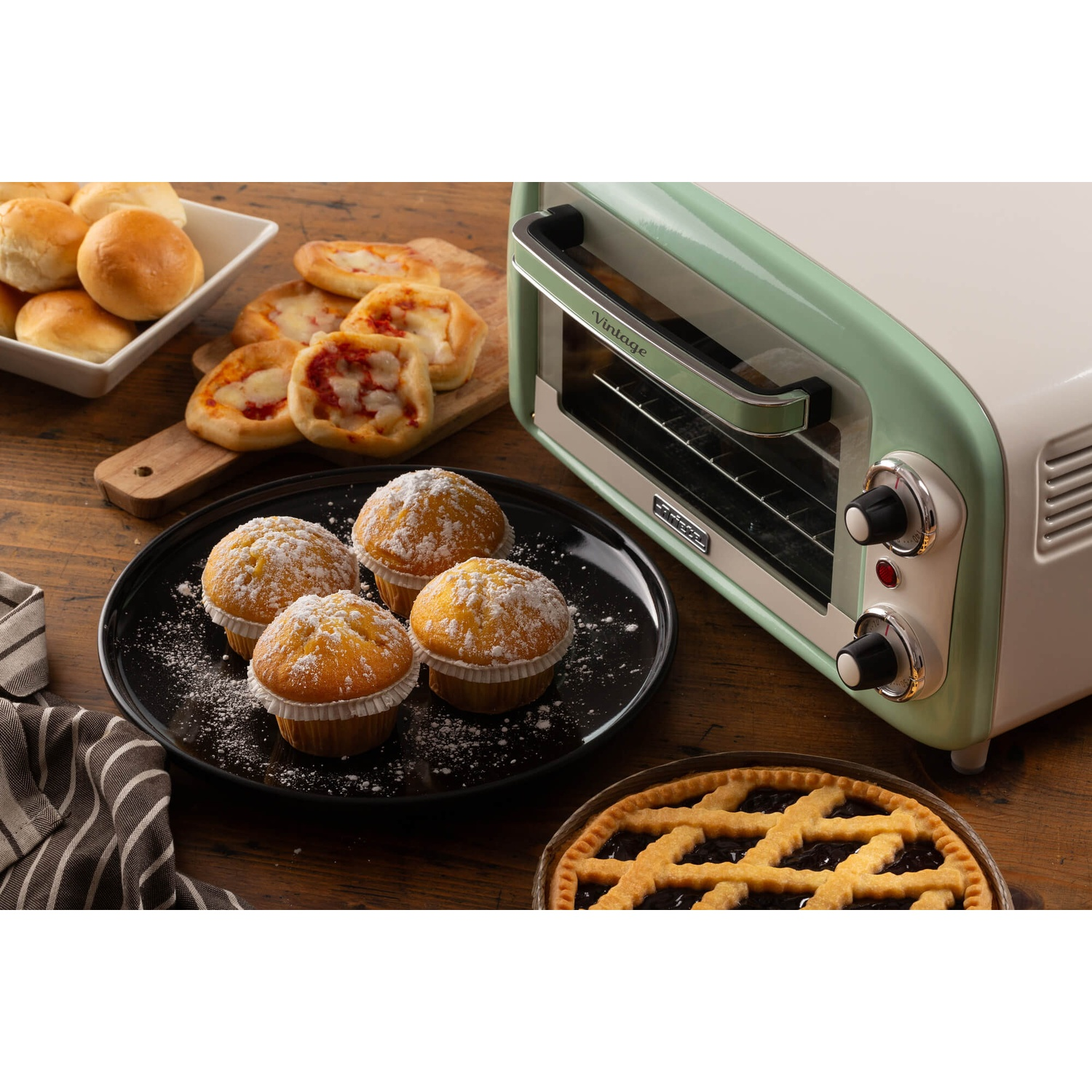 Un tostapane in stile vintage con prodotti da forno: muffin, mini pizze, panini e una torta su un tavolo di legno.